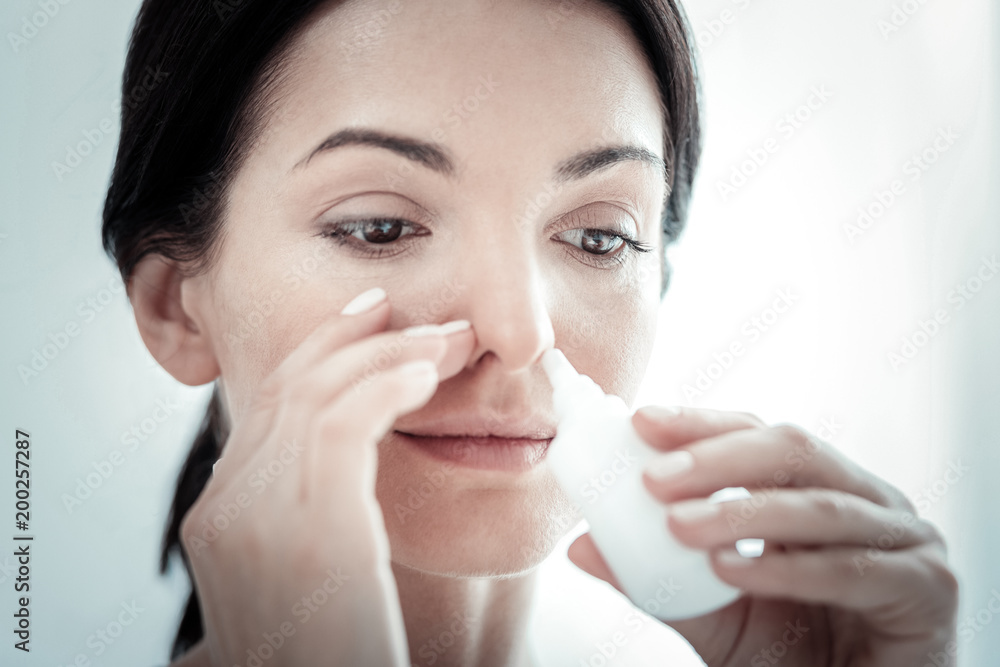 Feel better myself. Pleasant satisfied ill woman spending time in the bright room smiling and using nose spray. Feel better myself. Pleasant satisfied ill woman spending time in the bright room smiling and using nose spray.