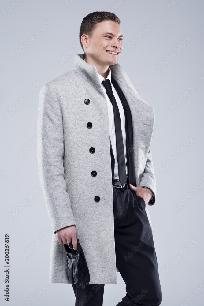 Fototapeta premium Handsome young man in a gray coat and black suit stands on a gray background