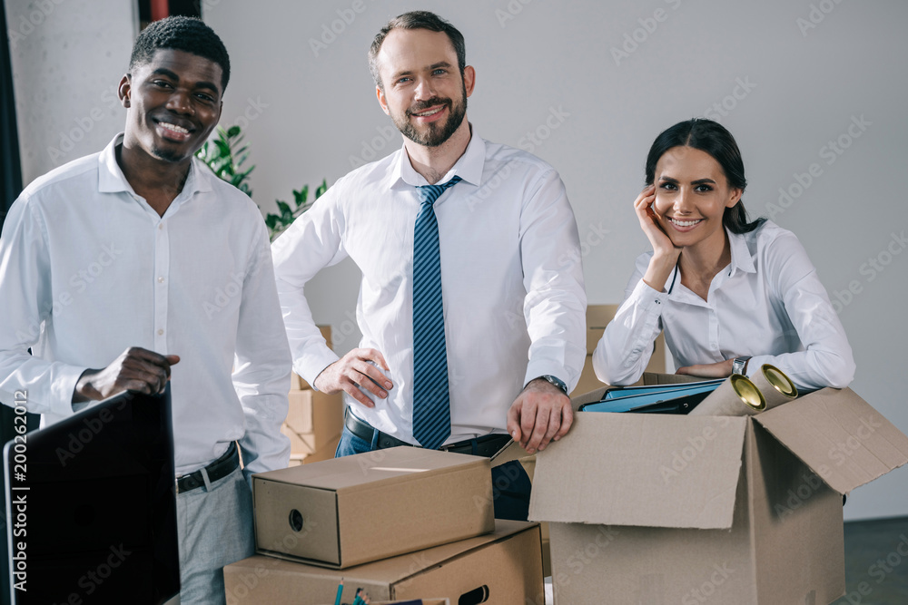 happy multiethnic colleagues smiling at camera while unpacking cardboard boxes in new office happy multiethnic colleagues smiling at camera while unpacking cardboard boxes in new office