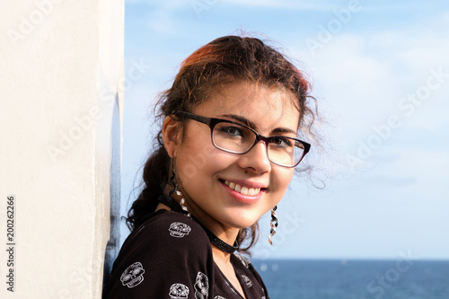 Portrait of an attractive and young woman (mixed ethnicities of Caucasian and Nepalese) wearing glasses and smiling at the ocean.