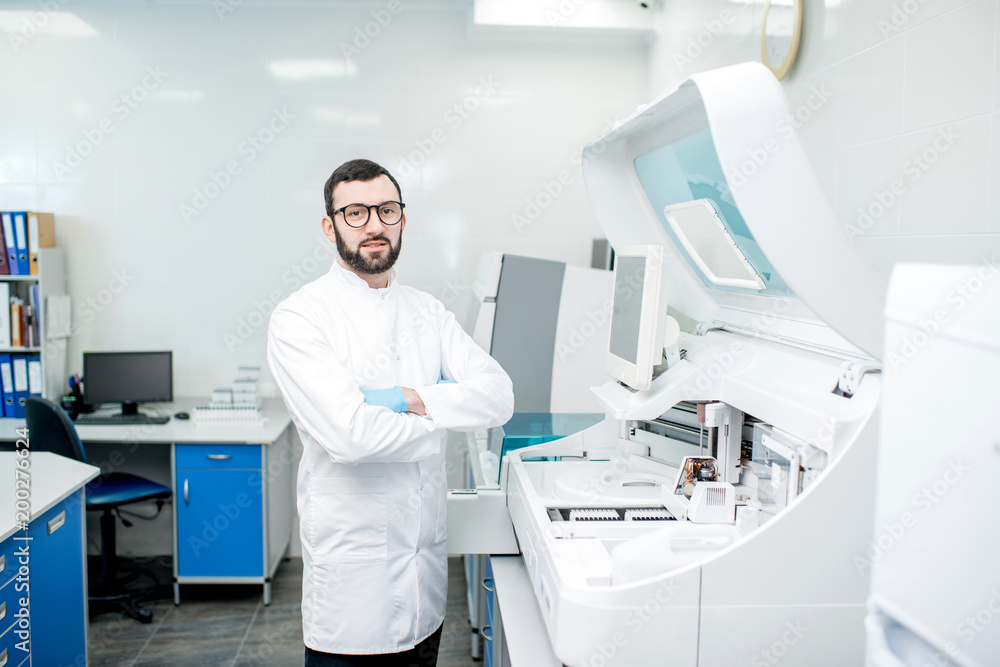 Portrait of a male laboratory assistant in uniform standing near the medical analizer machine indoors Portrait of a male laboratory assistant in uniform standing near the medical analizer machine indoors