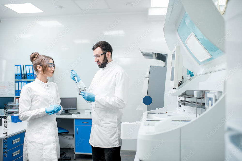 Couple of laboratory assistants in uniform working with test tubes standing near the analyzer machine in the laboratory Couple of laboratory assistants in uniform working with test tubes standing near the analyzer machine in the laboratory