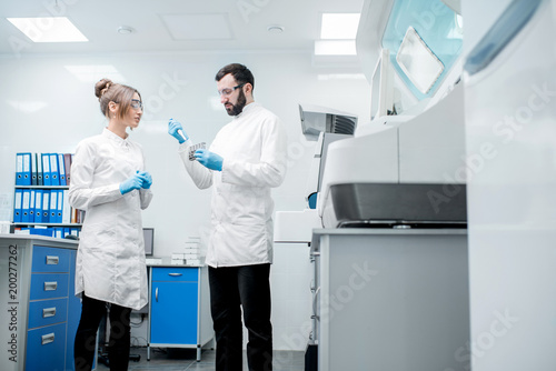 Couple of laboratory assistants in uniform working with test tubes standing near the analyzer machine in the laboratory