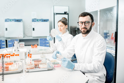 Portrait of a man in medical uniform during the bacteriological tests sitting with assistant in the modern laboratory