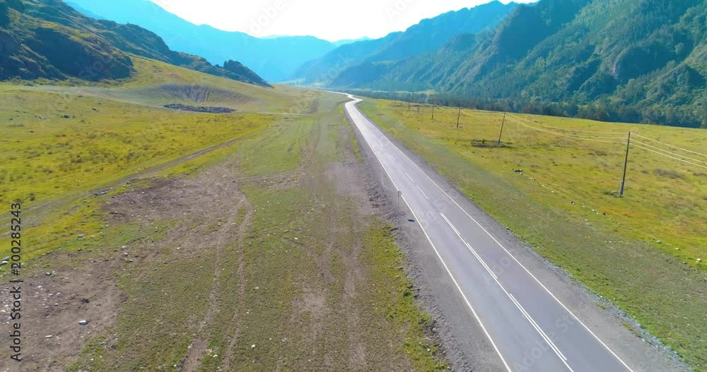 Aerial UHD 4K view. Low air flight over mountain asphalt highway road and meadow at sunny summer morning. Near green trees. Fast horizontal movement with sun rays. Aerial UHD 4K view. Low air flight over mountain asphalt highway road and meadow at sunny summer morning. Near green trees. Fast horizontal movement with sun rays.