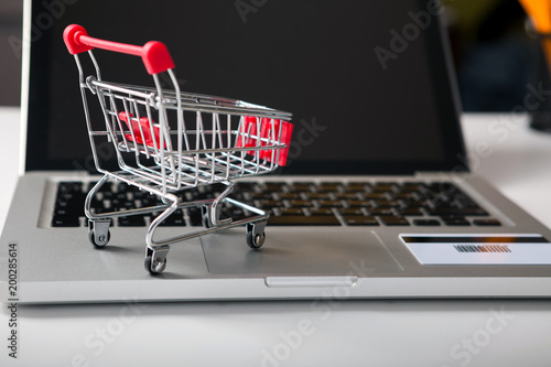 Online shopping. Bank card nearby a laptop and mini shopping cart on white background top view.