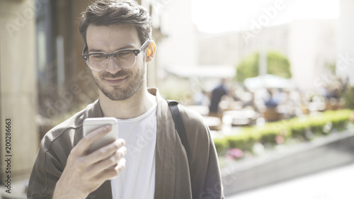 A young smiling man wearing glasses scrolls the phone in the street. A smiling young man dressed in casual scrolling his cellphone outdoors
