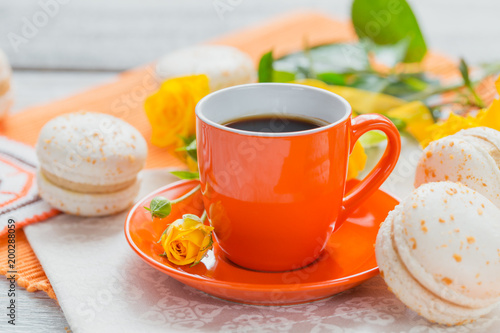 Orange cup of black coffee, yellow roses flowers and sweet pastel french macaroons