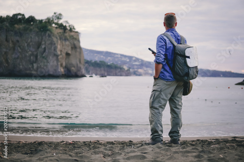 man, guy the photographer working look at the phone, receives a call, calls on the beach, backpack, concept of find information at travel to foregin country, communication, mountains on background