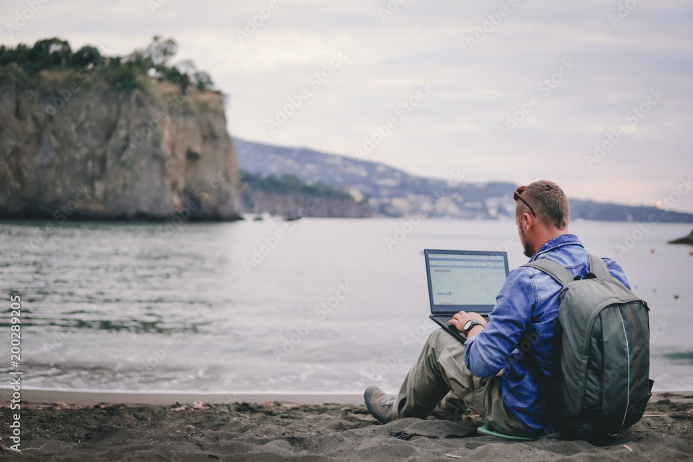 man, guy the photographer working with a laptop on the beach, backpack, concept of find information at travel to foregin country, communication, mountains on background