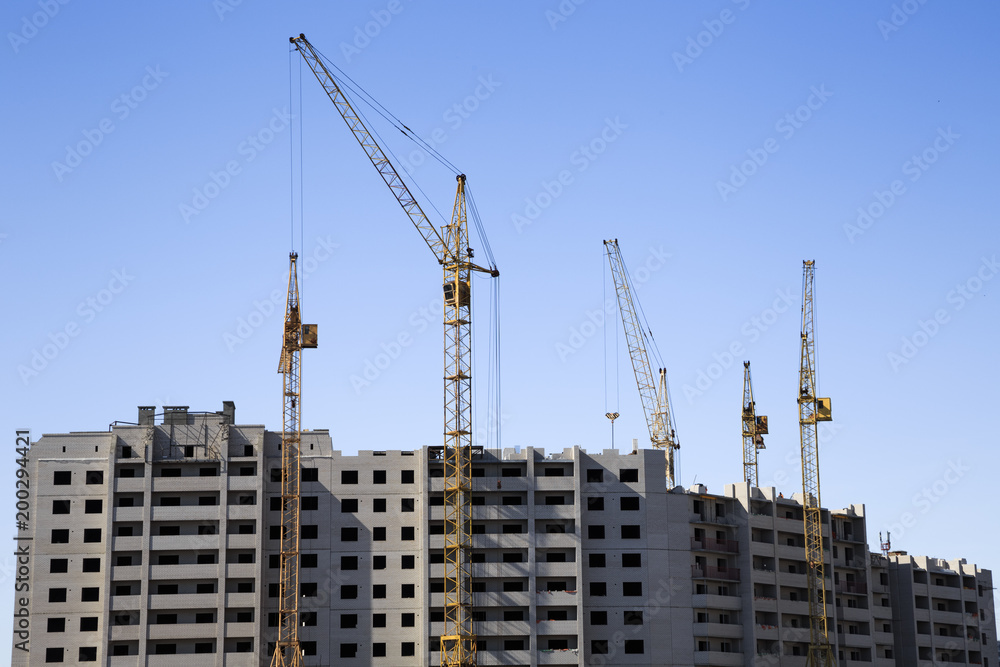 Distant view of a construction site with an unfinished apartment building and several cranes. Clear blue sky serves as a background. Distant view of a construction site with an unfinished apartment building and several cranes. Clear blue sky serves as a background.