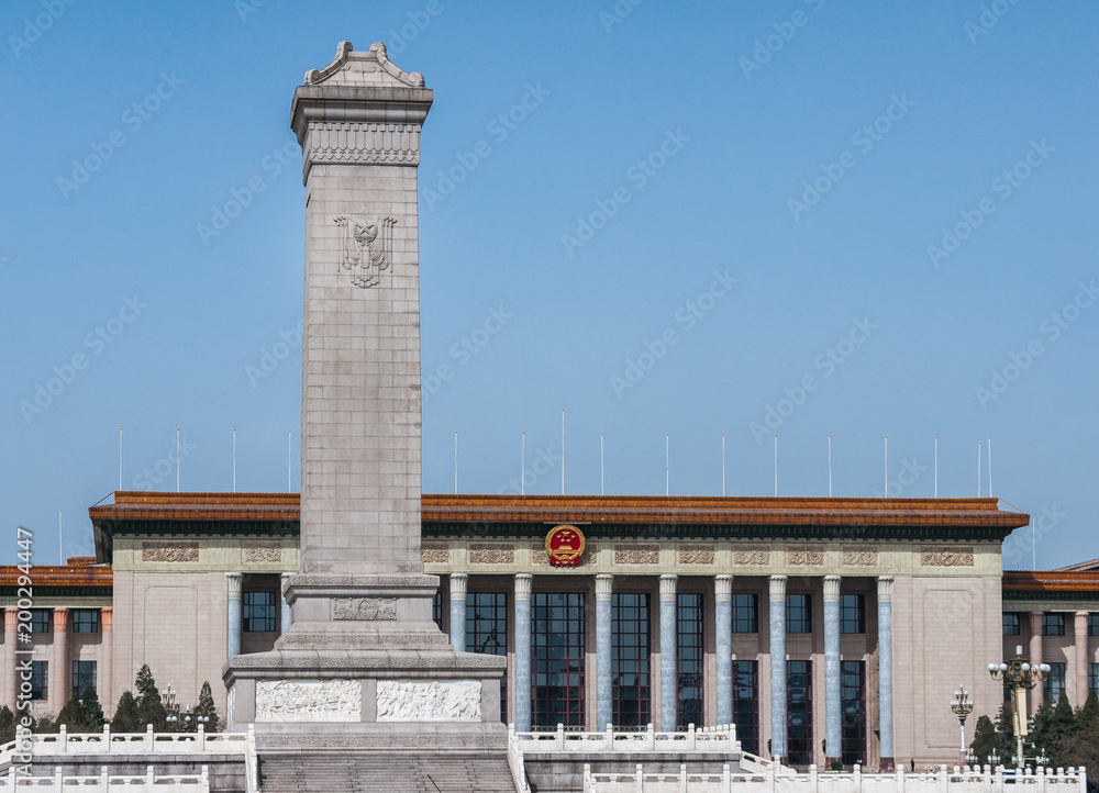 Beijing, China - April 27, 2010: Beige tall square obelisk of war memorial to the People’s Heroes at Tienanmen Square against ligh blue sky. Back is facade of Great Hall of the People.
