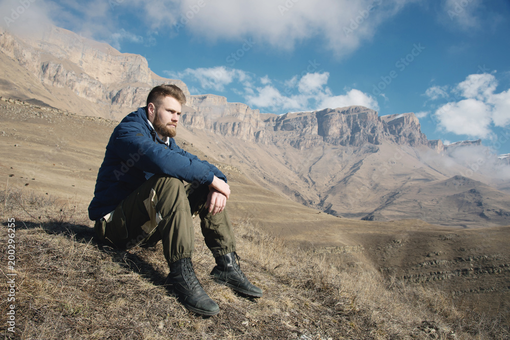 A portrait of a stylish bearded hipster sitting on a rock against the backdrop of epic rocks and contemplating into the distance thinking about life. The concept of tranquility and solitude with A portrait of a stylish bearded hipster sitting on a rock against the backdrop of epic rocks and contemplating into the distance thinking about life. The concept of tranquility and solitude with