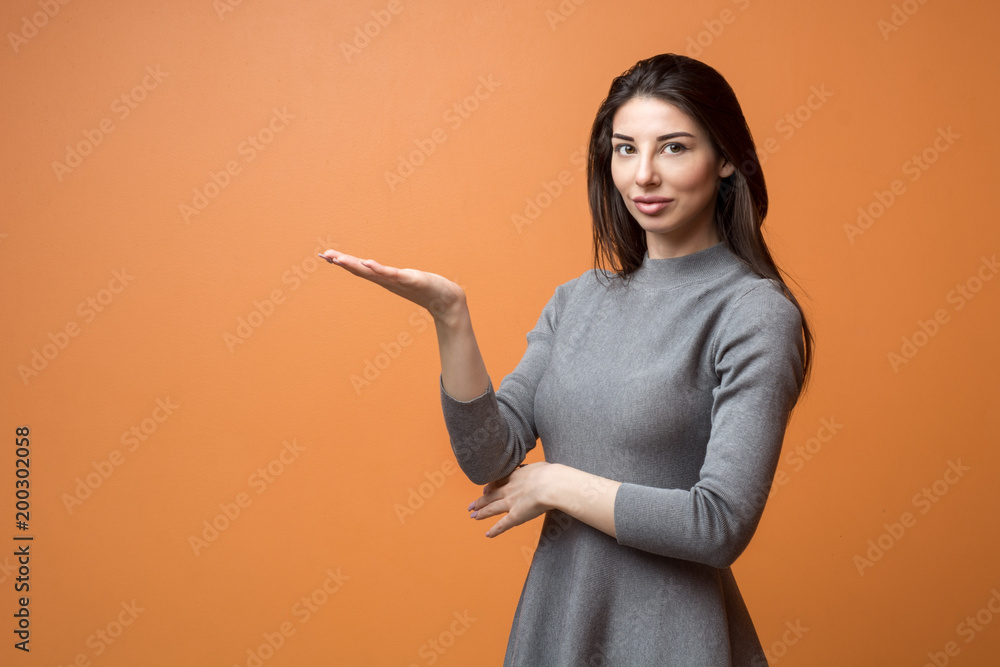 Portrait of a young beautiful business woman in neutral dress holding out empty hand and looking into camera isolated on a orange background Portrait of a young beautiful business woman in neutral dress holding out empty hand and looking into camera isolated on a orange background