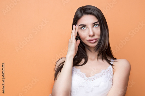 Closeup portrait of a young beautiful brunette pensive woman holding her hand new her head and looking up, think about something against orange background
