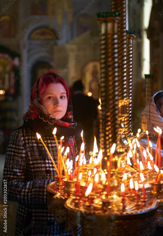 Praying young woman with candle near pedestal with many other candles in church by her hand to show her faith and esteem to God Praying young woman with candle near pedestal with many other candles in church by her hand to show her faith and esteem to God