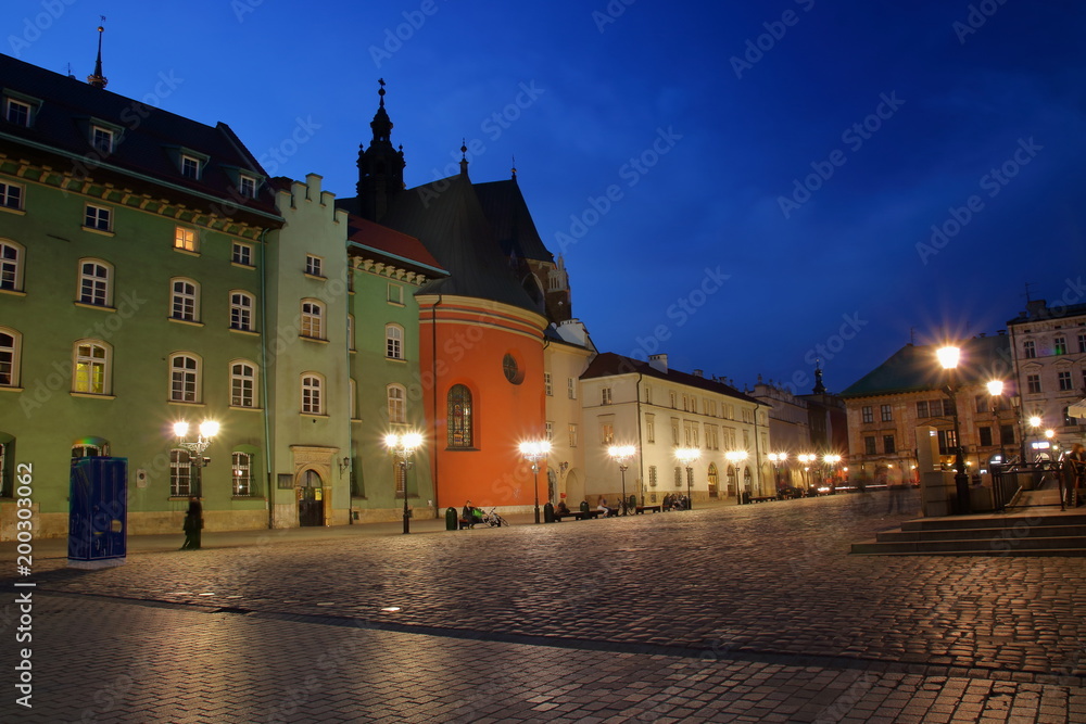 Nocny widok Małego Rynku w Krakowie, Polska, zabytkowa architektura, ludzie odpoczywający na ławkach, uliczne lampy, ciemnogranatowe niebo