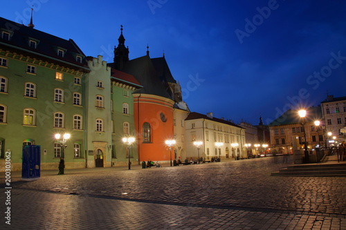 Nocny widok Małego Rynku w Krakowie, Polska, zabytkowa architektura, ludzie odpoczywający na ławkach, uliczne lampy, ciemnogranatowe niebo