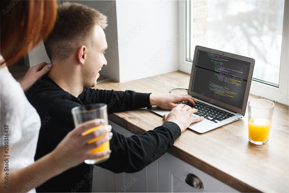 The wife hugs her husband in the kitchen. The husband sits at the computer and works, programist The wife hugs her husband in the kitchen. The husband sits at the computer and works, programist