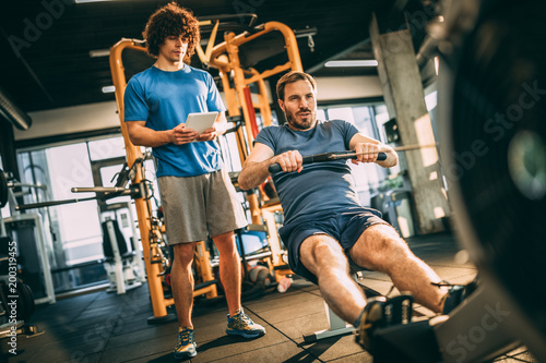Young man with his personal instructor in fitness center. Using rowing machine for cardio training in the gym.