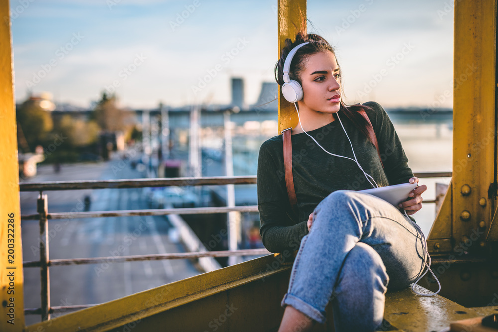 Young talented female student dressed in casual clothing sits on the top of the building and listening to favorite radio station via earphones.