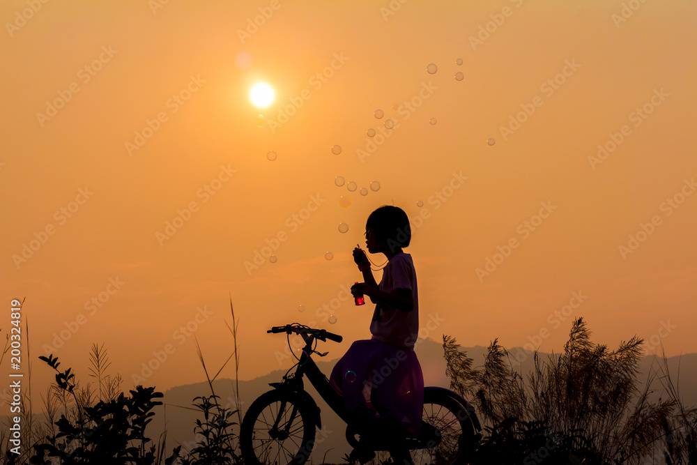 Fototapeta premium Silhouette little girl sitting on bicycle playing with bubble wand on mountain at sunset background