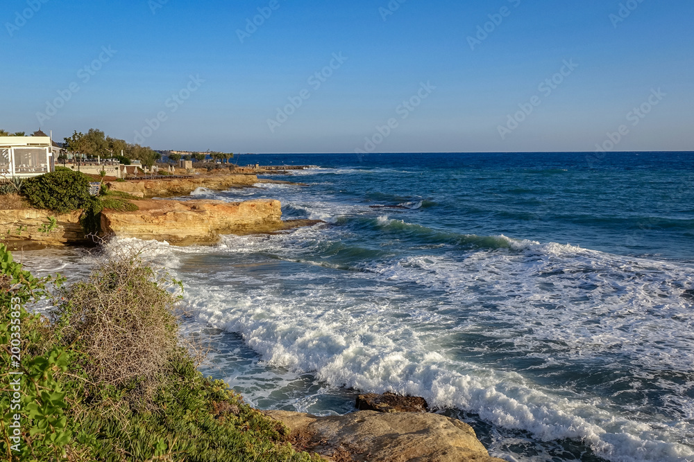 Mediterranean rocky coastline, blue cloudless clouds, blue sea with white waves off the coast, Crete, Greece.