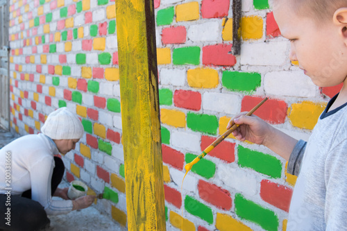 Happy caucasian child paints a brick wall with paint brush in different colors. Red, blue, yellow, green. Capacity with paint and hands close-up view