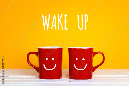 Two red coffee mugs with a smiling faces on a yellow background with the phrase Wake up.