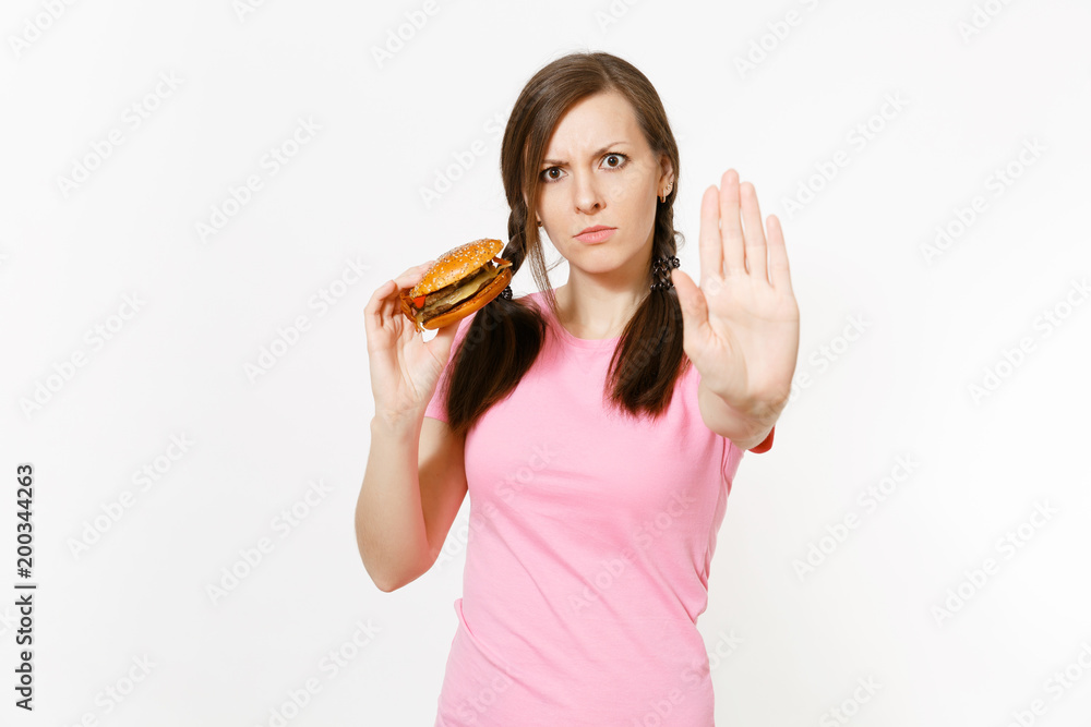 Young strict woman showing stop gesture with palm, holding in hands burger isolated on white background. Proper nutrition or American classic fast food. Copy space for advertisement. Advertising area. Young strict woman showing stop gesture with palm, holding in hands burger isolated on white background. Proper nutrition or American classic fast food. Copy space for advertisement. Advertising area.