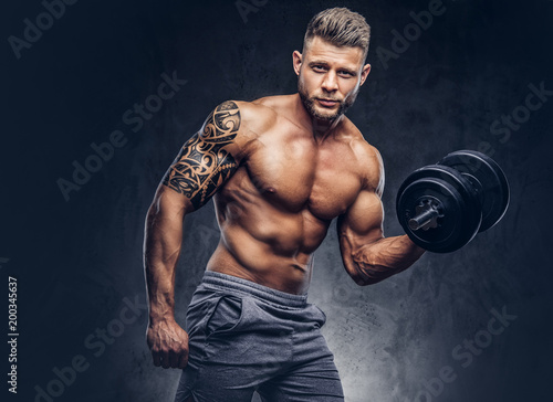 A handsome shirtless tattooed bodybuilder with stylish haircut and beard, wearing sports shorts, posing in a studio. Isolated on a dark background © Fxquadro
