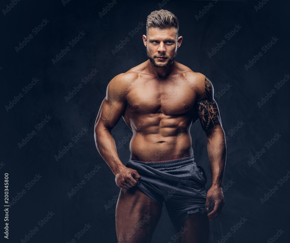 Fototapeta premium A handsome shirtless tattooed bodybuilder with stylish haircut and beard, wearing sports shorts, posing in a studio. Isolated on a dark background