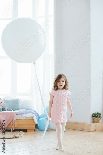 Cute little girl in pink dress walking with white balloon in the nursery. Portrait of a little child surprised