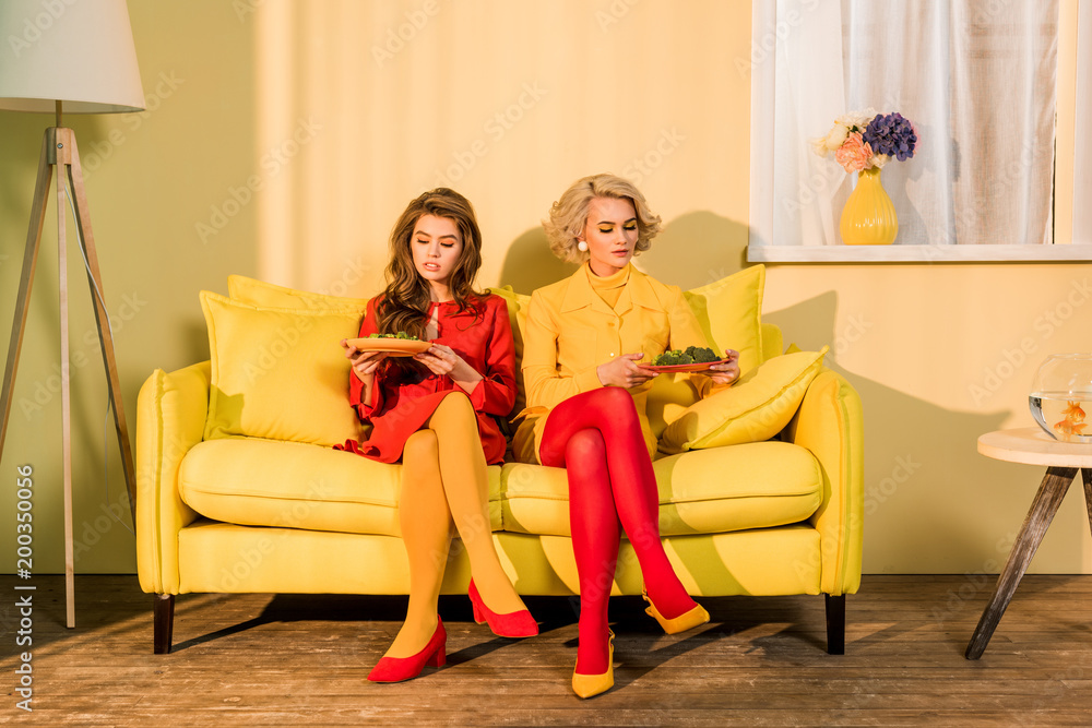 pretty women in retro clothing with vegetables on plates sitting on yellow sofa at bright room, doll house concept
