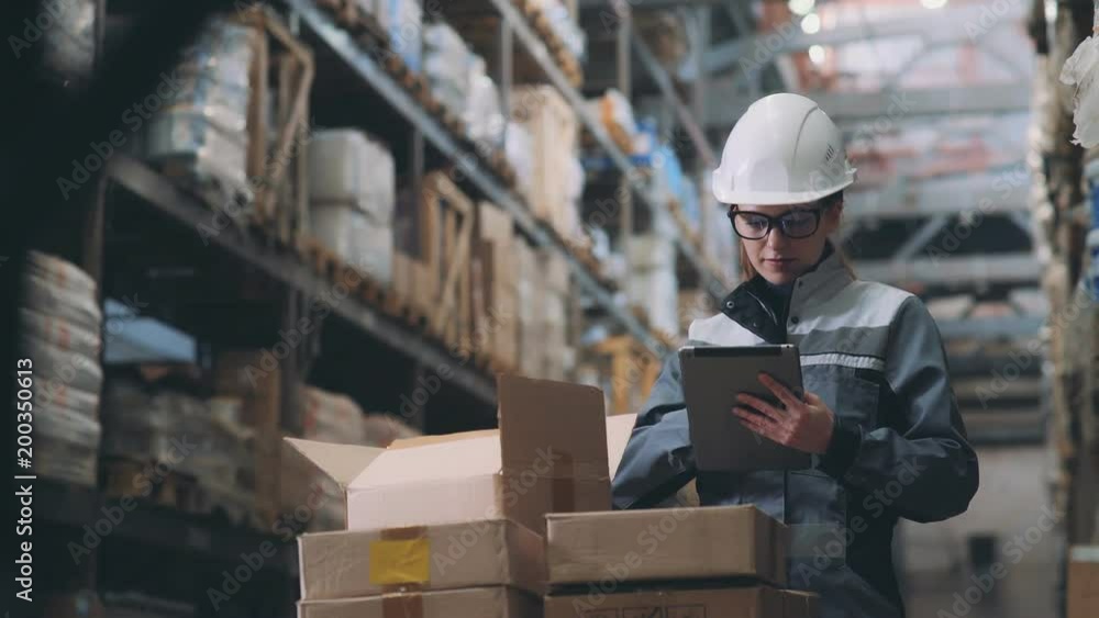 A woman in a warehouse in a white helmet and gray uniform opens a cardboard box, looks at the goods and makes an inventory. Merchandiser recounts the item in the box and makes notes on the tablet. A woman in a warehouse in a white helmet and gray uniform opens a cardboard box, looks at the goods and makes an inventory. Merchandiser recounts the item in the box and makes notes on the tablet.
