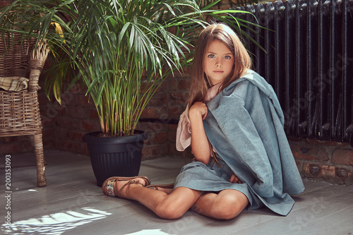 Cute little girl with long brown hair wearing a stylish dress, sitting on a wooden floor in a room with a loft interior