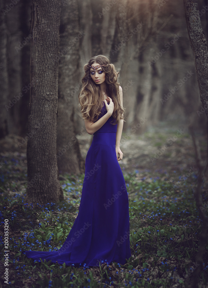 Girl elf with long hair and blue eyes in tiara and a long blue dress with a train walking through the spring blossoming forest. Girl Princess dreams. Girl elf with long hair and blue eyes in tiara and a long blue dress with a train walking through the spring blossoming forest. Girl Princess dreams.