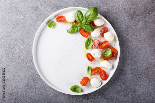 White plate of healthy classic delicious caprese salad with ripe tomatoes and mozzarella cheese with fresh basil leaves on gray concrete background with space for text. Italian food. Top view. © Iryna Melnyk