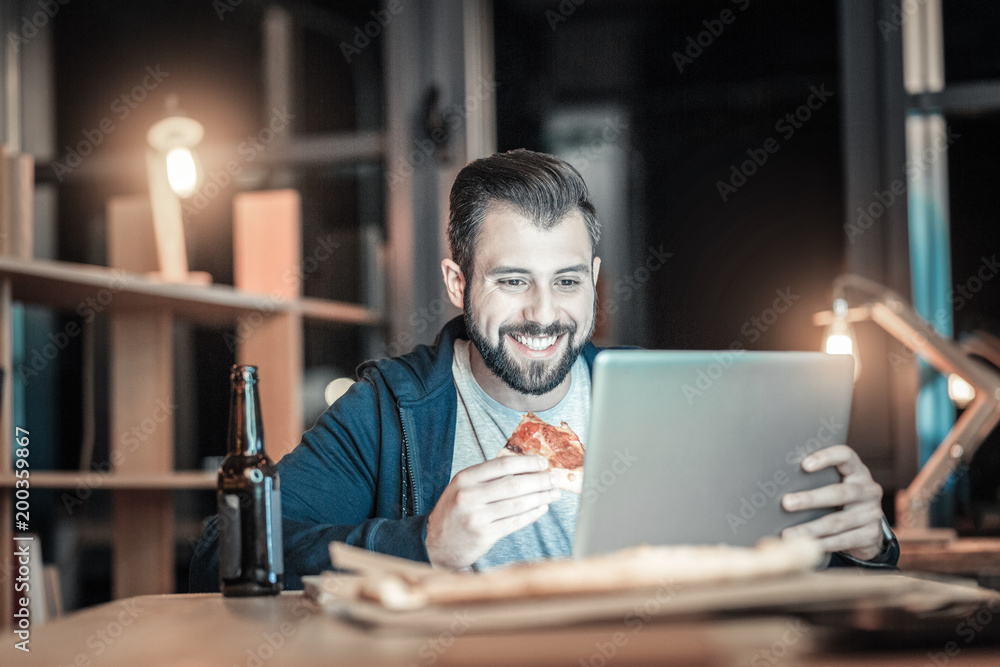 Pizza time. Delighted positive IT guy eating pizza while looking down and smiling Pizza time. Delighted positive IT guy eating pizza while looking down and smiling