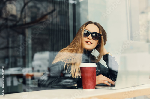 Young girl sit in coffee place in front of the window look at her laptop and drink tea from red cup