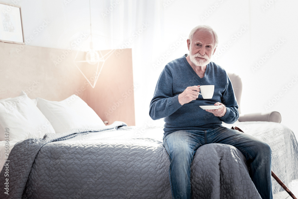 Tea time. Nice positive aged man sitting on the bed and having tea while resting at home