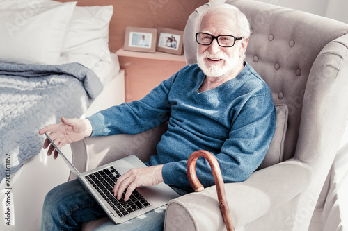 Digital technology. Happy bearded aged man sitting in the armchair and smiling while using a laptop