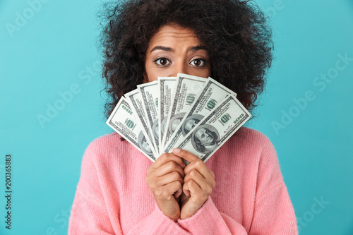 Portrait of excited woman 20s with afro hairstyle covering face with fan of money dollar bills, isolated over blue background