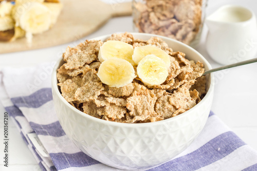 Whole wheat cornflakes with banana slices in white bowl, ingredients in the background.