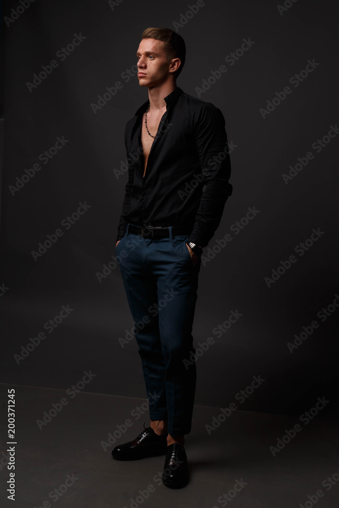 attractive white man in black shirt and blue pants stands on a dark background