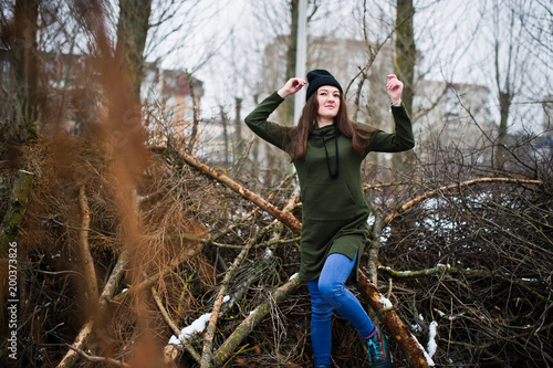 Young girl wear on long green sweatshirt, jeans and black headwear at branches of the pine tree in winter day.