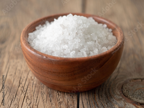 White bowl with large sea salt on brown bright dark wooden table. Top view, close up, selective focus