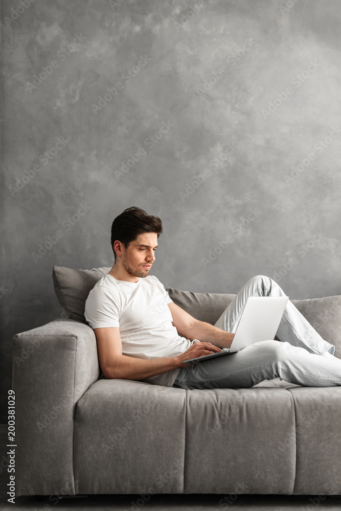 Attractive serious man 30s in basic clothing using laptop, while lying on couch in gray interior