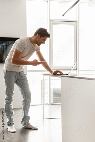 Full length photo in profile of attractive guy in basic clothing using mobile phone, and laptop at home