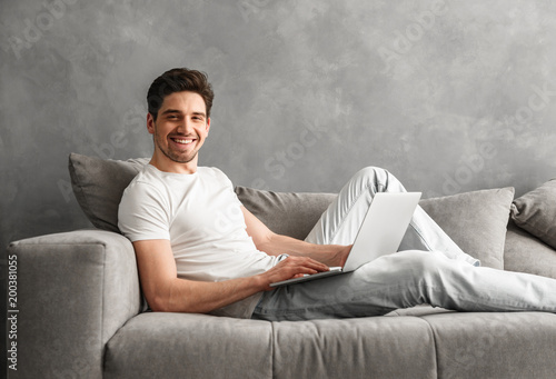 Caucasian unshaved man 30s in casual clothing using notebook, while lying on cozy sofa in gray apartment